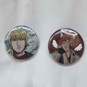 May include: Two round buttons with anime-style illustrations. One features a blonde-haired character with the text "Huh? wuh" on a blue and green background. The other button has a character with orange hair, a halo, and angel wings on a maroon background.