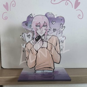 May include: An acrylic standee featuring an illustrated character with light pink hair wearing a peach-colored sweater. The character holds a black smartphone. The background includes purple heart shapes and a rectangular base with a purple gradient design.