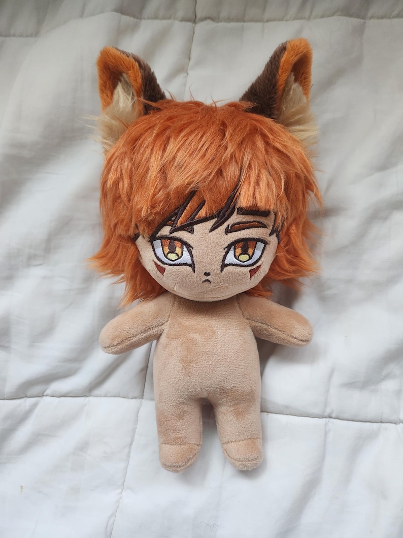 May include: A plush doll with orange hair and fox-like ears. The doll has a tan body, detailed facial features, and large, expressive eyes. The doll's hair is styled in a shaggy, layered cut. The ears are a mix of brown, orange, and tan.