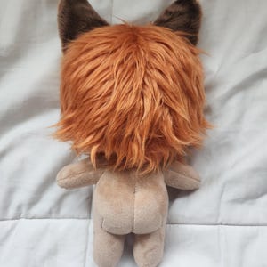 May include: A plush toy with a tan body and a head covered in orange and brown faux fur, resembling a fox or lion. The toy has small, brown ears and a simple, rounded body shape. The back view is shown.