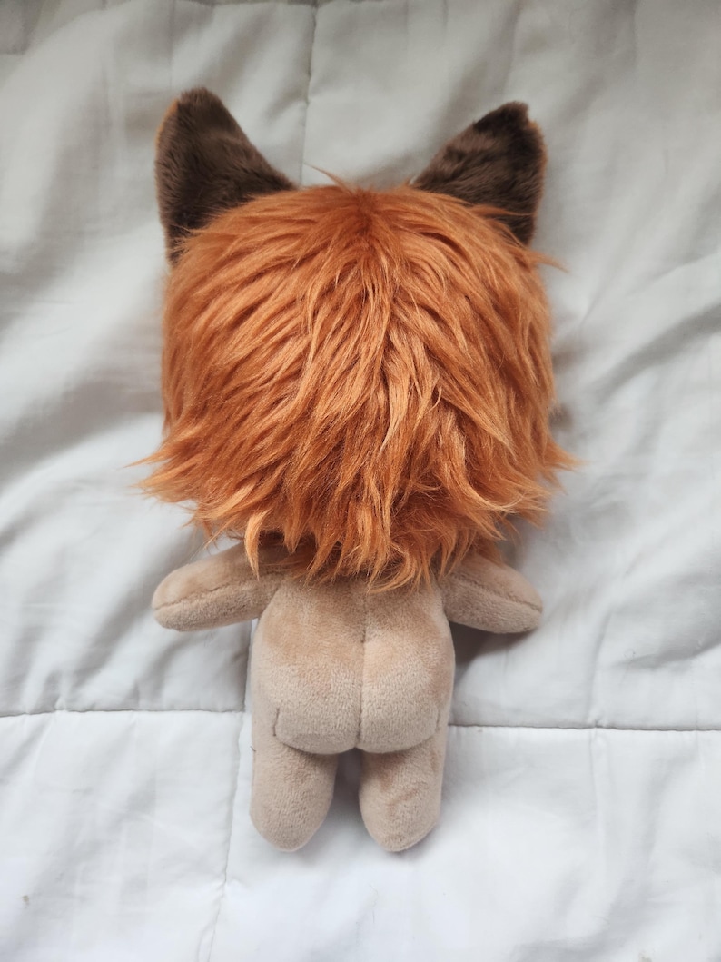 May include: A plush toy with a tan body and a head covered in orange and brown faux fur, resembling a fox or lion. The toy has small, brown ears and a simple, rounded body shape. The back view is shown.