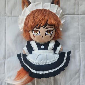 May include: A plush doll of a fox character wearing a black and white maid outfit, complete with a ruffled apron and a white cap. The doll has orange hair, fox ears, and a fluffy orange tail. The doll's eyes are large and detailed.