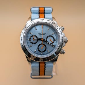 Puede incluir: Un reloj de pulsera de acero inoxidable con esfera azul claro y correa a rayas azul claro y naranja. La esfera del reloj tiene tres subesferas y el texto "Hands-on Time". El reloj tiene un bisel plateado con marcas negras.