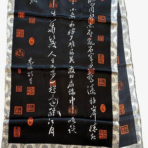 Può includere: Sciarpa in seta nera con calligrafia bianca e sigilli arancioni. Presenta un bordo argentato e grigio a motivi. La calligrafia è disposta verticalmente sullo sfondo nero.