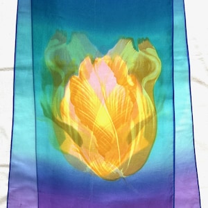 Op de afbeelding: Een doorschijnende sjaal met een bloemenontwerp. De sjaal heeft een verloop van blauw, blauwgroen en paars. Een grote, gestileerde gele en oranje tulp is het middelpunt. De sjaal heeft een donkerblauwe rand.