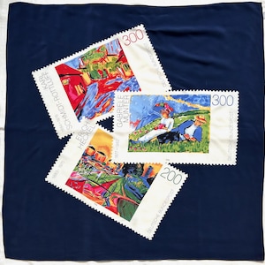 May include: Three postage stamps with vibrant artwork are arranged on a dark blue fabric. The stamps showcase paintings by Karl Schmidt-Rottluff, Erich Heckel, and Gabriele Münter. Each stamp has a value of 200 or 300, with the text "DEUTSCHE BUNDESPOST".
