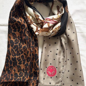 Könnte beinhalten: Seidenschal mit verschiedenen Mustern. Eine Seite zeigt einen Leopardenprint in Braun und Schwarz. Eine andere Seite hat einen beigen Hintergrund mit schwarzen Sternen und einem rosa Logo. Die Mittelbahn hat einen cremefarbenen Hintergrund mit Reitmotiven.