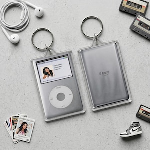 May include: Silver iPod keychain with a clear protective case, displaying a music track. The back of the keychain has the name "Doey". Additional items include a pair of white earbuds, cassette tapes, a miniature sneaker, and a stack of cards.