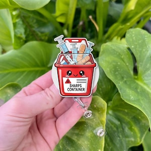 May include: A red sharps container badge reel with a cartoon design. The container is filled with syringes and bandages. The badge reel has the words "SHARPS CONTAINER" and a biohazard symbol.