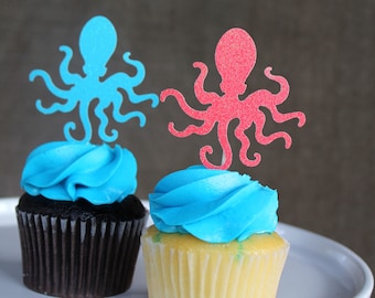 Puede incluir: Dos cupcakes decorados con glaseado azul y coronados con un recorte de pulpo. Un pulpo es azul y el otro es rosa.