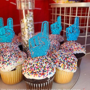 May include: Blue foam finger cupcake toppers with glitter. The toppers are on cupcakes with colorful sprinkles.