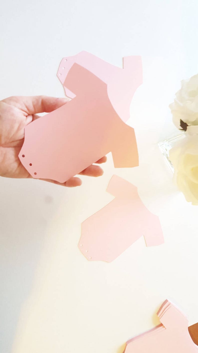 Op de afbeelding: Roze papieren uitsnijdingen in de vorm van baby-rompers. De rompers zijn plat en hebben kleine gaatjes die in de bovenste hoeken zijn geponst.
