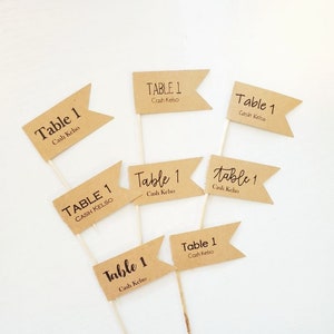 May include: A set of six brown paper table number flags with the text "Table 1" and "Cash Kelso" printed on each flag. The flags are attached to wooden skewers.