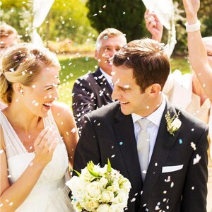 Puede incluir: Una novia y un novio caminan por el pasillo, sonriendo y riendo mientras cae confeti a su alrededor. La novia lleva un vestido de novia blanco y el novio lleva un traje negro con camisa blanca y corbata plateada. El novio lleva un ramo de flores blancas.