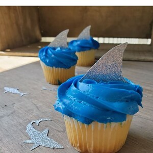 May include: Blue and white cupcakes decorated with silver glitter shark fin toppers. The cupcakes are on a wooden surface with silver glitter shark cutouts.