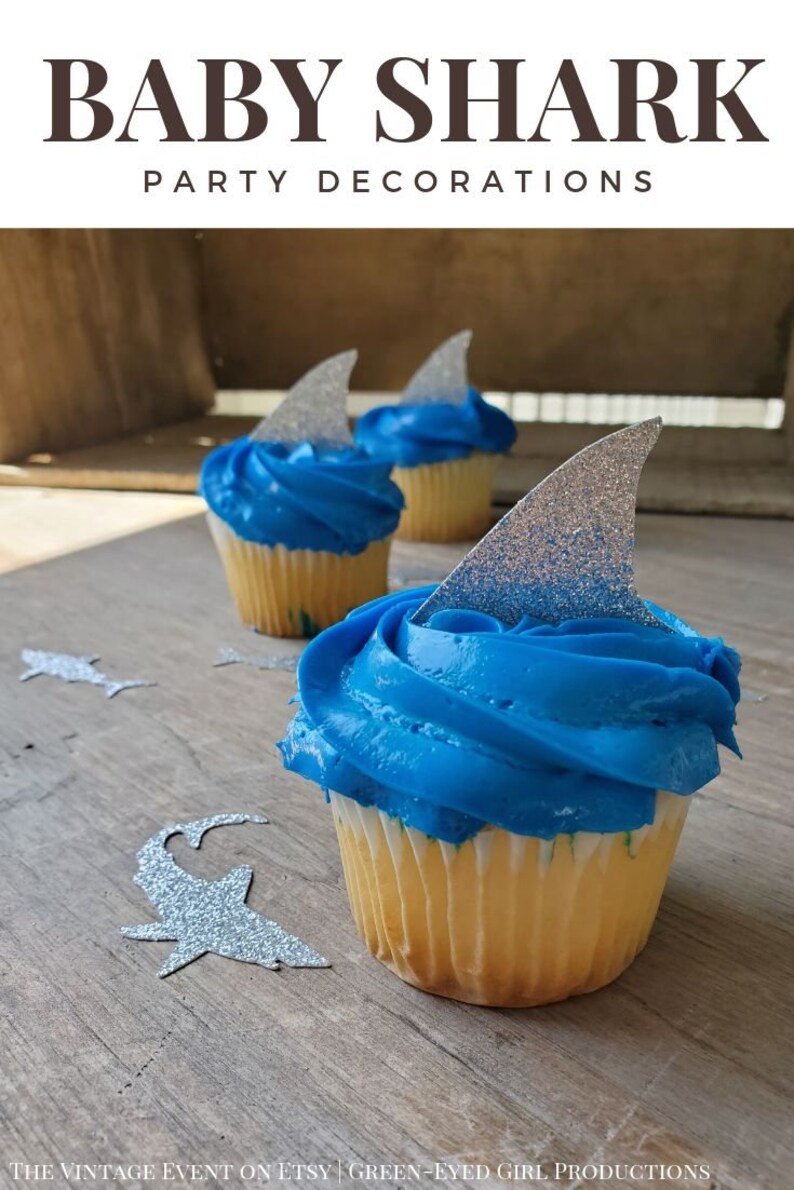 May include: Blue and white cupcakes decorated with silver glitter shark fin toppers. The cupcakes are on a wooden surface with silver glitter shark cutouts.