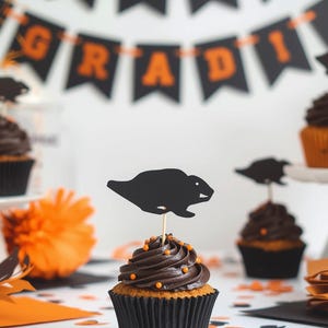 Puede incluir: Un cupcake decorado con glaseado de chocolate y chispas naranjas, coronado con una silueta negra de castor. El cupcake forma parte de una exhibición temática de graduación, con una pancarta que dice "BODY GRADI".