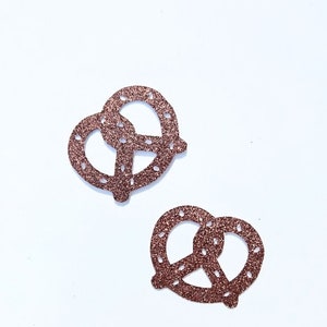 May include: Two gold glitter pretzel confetti pieces on a white background. The confetti is shaped like pretzels.