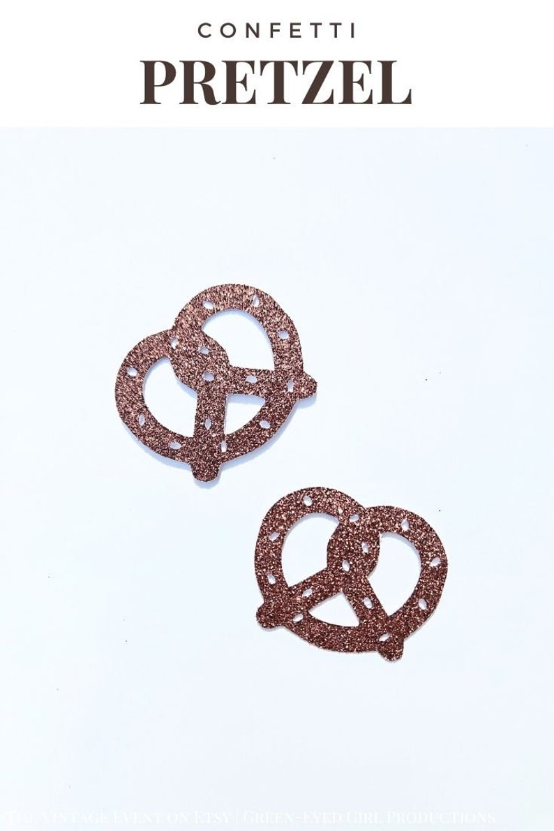 May include: Two gold glitter pretzel confetti pieces on a white background. The confetti is shaped like pretzels.