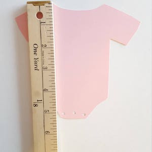 Op de afbeelding: Een roze papieren uitsnede van een rompertje met drie kleine gaatjes aan de onderkant. Het rompertje is 12,7 cm hoog en wordt gemeten tegen een houten liniaal.