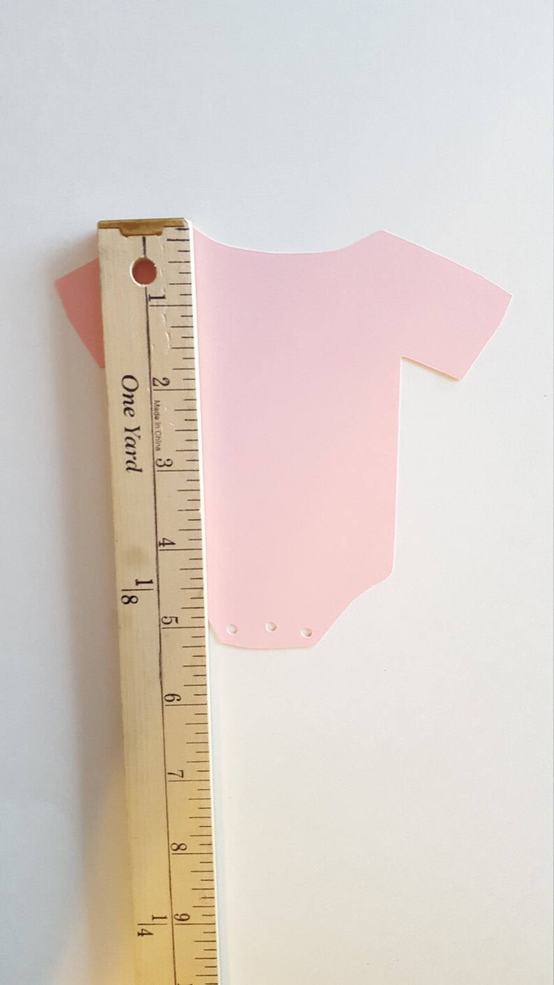 Op de afbeelding: Een roze papieren uitsnede van een rompertje met drie kleine gaatjes aan de onderkant. Het rompertje is 12,7 cm hoog en wordt gemeten tegen een houten liniaal.