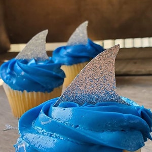 May include: Three cupcakes with blue frosting and silver glitter shark fin toppers. The cupcakes are on a wooden surface.  CUPCAKE TOPPERS SHARK