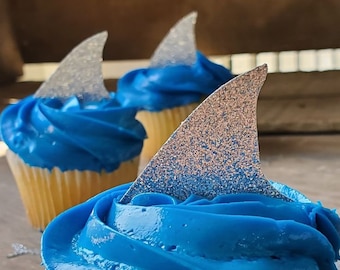 May include: Three cupcakes with blue frosting and silver glitter shark fin toppers. The cupcakes are on a wooden surface.  CUPCAKE TOPPERS SHARK