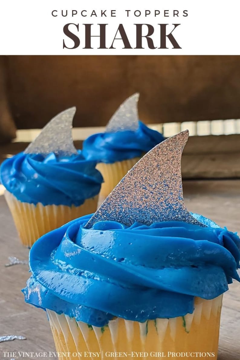 May include: Three cupcakes with blue frosting and silver glitter shark fin toppers. The cupcakes are on a wooden surface.  CUPCAKE TOPPERS SHARK