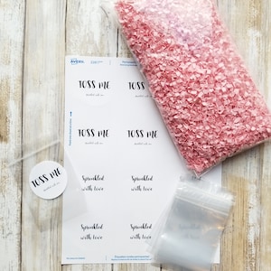Puede incluir: Una hoja de papel blanco con etiquetas que dicen "Toss Me" y "Sprinkled with Love". Tambi&eacute;n hay varias bolsas de pl&aacute;stico transparentes y una bolsa de confeti rosa.