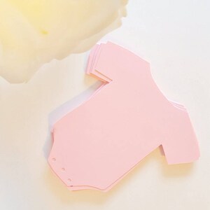 Op de afbeelding: Een stapel roze papieren uitsnijdingen in de vorm van baby-rompers.