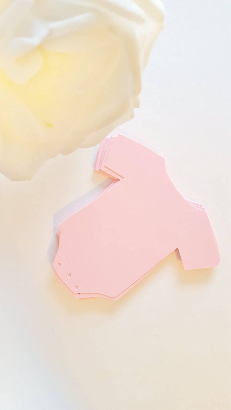 Op de afbeelding: Een stapel roze papieren uitsnijdingen in de vorm van baby-rompers.