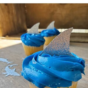 May include: Blue and white cupcakes with silver glitter shark fin toppers.  "Baby Shark Party Decorations" is written in black text at the top of the image.