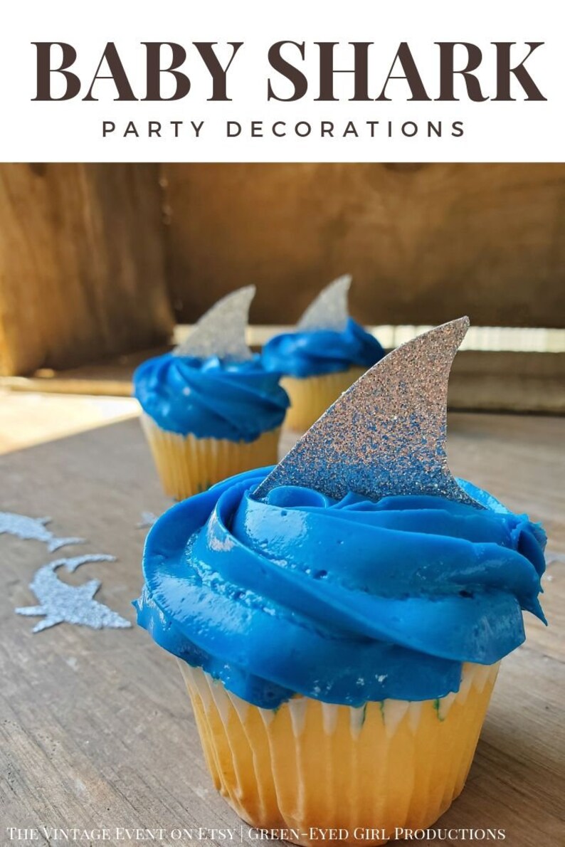 May include: Blue and white cupcakes with silver glitter shark fin toppers.  "Baby Shark Party Decorations" is written in black text at the top of the image.