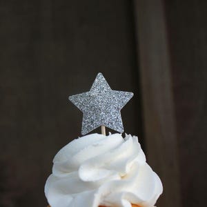 May include: Silver glitter star cupcake topper. The star is on top of a white frosted cupcake.