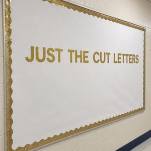 May include: A rectangular bulletin board with a white background and a scalloped gold glitter border. The text JUST THE CUT LETTERS is displayed in the center in gold glitter capital letters. The board is mounted on a light-colored wall.