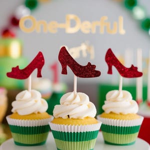 May include: Three cupcakes with white icing and red glitter high heel decorations. The cupcakes are on a white cake stand. The cupcake cases are green and white. The background has the text "One-Derful".