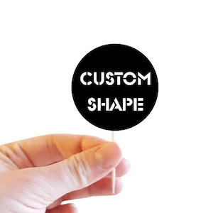 Puede incluir: Un adorno para cupcakes con el texto "CUSTOM CUT" en la parte superior y un círculo negro con "CUSTOM SHAPE" en blanco. El adorno lo sostiene una mano sobre un fondo blanco. También se ve el texto "CUPCAKE TOPPERS".