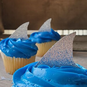 May include: Three blue frosted cupcakes with silver glitter shark fin toppers. The cupcakes are on a wooden surface.  CUPCAKE TOPPERS SHARK THE VINTAGE EVENT ON ETSY GREEN-EYED GIRL PRODUCTIONS