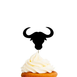 May include: Black silhouette of a Highland cow head cupcake topper. The topper is on a white cupcake with white frosting.