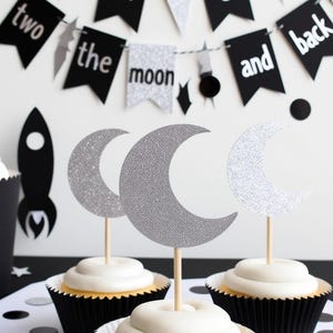 May include: A festive dessert display featuring cupcakes with white frosting and silver glitter moon toppers. A banner reads "two the moon and back" with a rocket ship and confetti, creating a space-themed party atmosphere.
