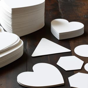 May include: A collection of white paper shapes, including hearts, a triangle, a circle, and hexagons, arranged on a dark brown wooden surface.