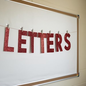 May include: Red glitter letters spelling "LETTERS" are strung across a white board. The letters are attached to a thin string with small silver clips. The board has a brown border and is mounted on a light beige wall.