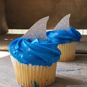 May include: Two cupcakes with blue frosting and silver glitter shark fin toppers.  The cupcakes are on a wooden surface with silver glitter shark cutouts.