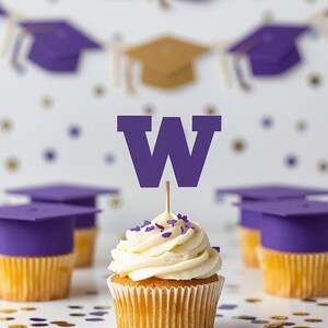 Puede incluir: Un cupcake con glaseado blanco y chispas moradas, coronado con una "W" morada sobre un palillo. Cupcakes con temática de graduación con adornos de birrete morado en el fondo, con una pancarta de birretes.
