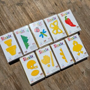 May include: A set of nine yellow die-cut craft shapes packaged in individual clear plastic sleeves. The shapes include a butterfly, a ladybug, a dragonfly, a chilli pepper, a rooster, a baseball and bat, a football and helmet, a flower, and a potted plant. Each package has the Sizzix logo and the words "Cots Originals" printed on it.