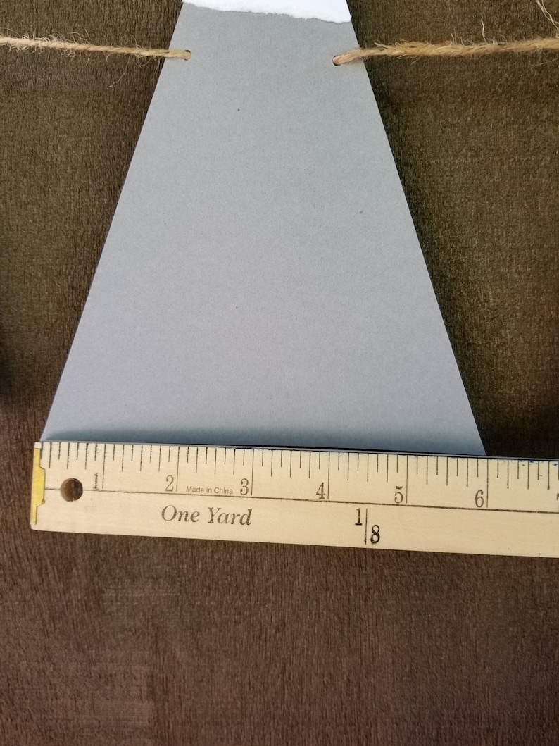 May include: A gray triangle-shaped paper banner with two holes punched at the top. The banner is hanging from a string. A wooden ruler with the words "One Yard" and "Made in China" printed on it is placed below the banner.