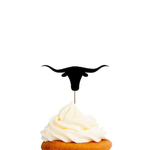 May include: A cupcake topped with a black silhouette of a longhorn bull. The cupcake is frosted with white frosting and has a yellow cupcake liner. The text "CUPCAKE TOPPERS LONGHORN" is at the top of the image.