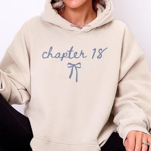 Op de afbeelding: Een zandkleurige hoodie met de tekst "chapter 18" in lichtblauw schrift, met een strik. De hoodie wordt gecombineerd met een zwarte legging en witte sneakers. Een drankje in een glas wordt in de hand gehouden.