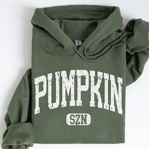 May include: A military green hooded sweatshirt featuring the white distressed text PUMPKIN SZN printed across the chest. The hoodie includes a drawstring hood and long sleeves, with the text PUMPKIN in a large arched font and SZN in a small box.