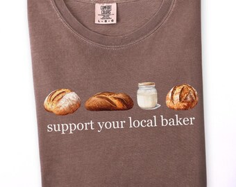 May include: Espresso-coloured t-shirt with a crew neck. The shirt displays a graphic of three loaves of bread and a jar of milk, with the text "support your local baker" in white. The shirt is folded.